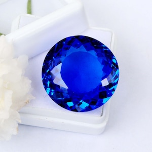 May include: A round, faceted gemstone with a deep, vibrant blue color. The stone features a brilliant cut with numerous triangular and kite-shaped facets that reflect light, creating a sparkling appearance. It is displayed in a white box.
