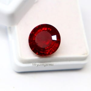 May include: A round, faceted, deep red gemstone with a brilliant cut, displayed on a white square jewellery box. The text "Shyamgems" is printed in black on the surface of the white box, positioned directly below the vibrant red gemstone.