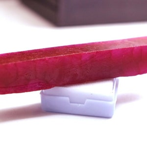 May include: A deep pink rectangular gemstone on a white display stand. The gemstone has a smooth, polished surface and a slightly translucent appearance.