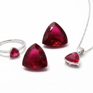 May include: A jewelry set consisting of a silver ring with a triangular red gemstone, two loose triangular red gemstones, and a silver pendant necklace featuring a matching triangular red gemstone, all displayed against a plain white background.