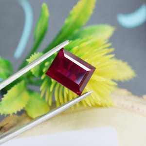May include: A deep red rectangular gemstone with a faceted cut. The gemstone is held by a pair of tweezers.
