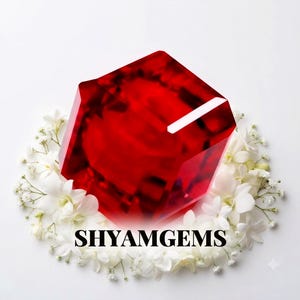 Könnte beinhalten: Ein großer, facettierter, tiefroter Edelstein in Würfelform, umgeben von einer kreisförmigen Anordnung kleiner weißer Blüten. Der Text SHYAMGEMS ist in schwarzen Großbuchstaben unten in der Mitte des Blumenarrangements aufgedruckt.