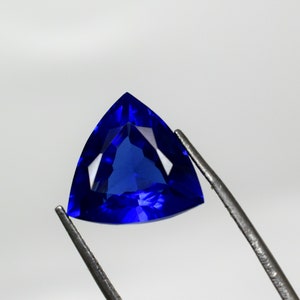 Può includere: Una pietra preziosa triangolare di colore blu intenso con sfaccettature, tenuta da una pinzetta su uno sfondo bianco.