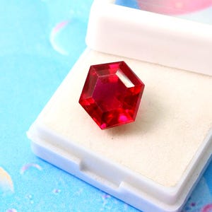 May include: A deep red hexagonal gemstone in a white jewelry box. The gemstone is faceted and has a high shine.