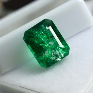 May include: A vibrant green emerald gemstone with a rectangular cut and a faceted finish. The emerald is set on a white background.