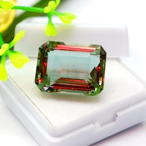 May include: A green and red gemstone with a rectangular emerald cut. The gemstone is displayed in a white box.