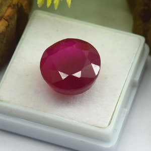 May include: A deep red gemstone with a faceted cut, displayed in a white box.
