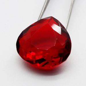Peut inclure: Une pierre précieuse rouge vif et profonde avec une taille en forme de cœur à facettes. La pierre présente une grande clarté et une réflexion de la lumière sur ses multiples surfaces polies, tenue par une pince métallique argentée.