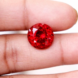 Pode incluir: Uma pedra preciosa redonda e facetada com uma cor vermelha vibrante e profunda. A pedra apresenta um corte brilhante com numerosas facetas triangulares e em forma de pipa que refletem a luz, criando um aspeto cintilante na superfície.