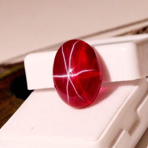 May include: A vibrant, oval-shaped, red gemstone with a star-like pattern of white lines. The gem is set on a white surface, showcasing its polished surface and internal light play. The gemstone is a striking example of natural beauty.