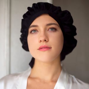 Peut inclure: Une femme portant un bonnet de nuit en satin noir. Le bonnet est froncé au sommet et a une longue cravate qui pend à l'arrière de son cou.