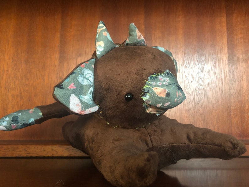 Op de afbeelding: Een bruin pluche speelgoed met een patchwork ontwerp. Het speelgoed heeft een lange staart en twee oren met een bloemenpatroon. Het speelgoed heeft een zwart knoopoog.