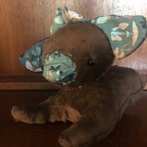 Op de afbeelding: Een bruin pluche speelgoed met een gestikt gezicht en stoffen oren. De oren zijn versierd met een groen, bruin en wit patroon. Het speelgoed heeft een gestikte neus en zwarte ogen.
