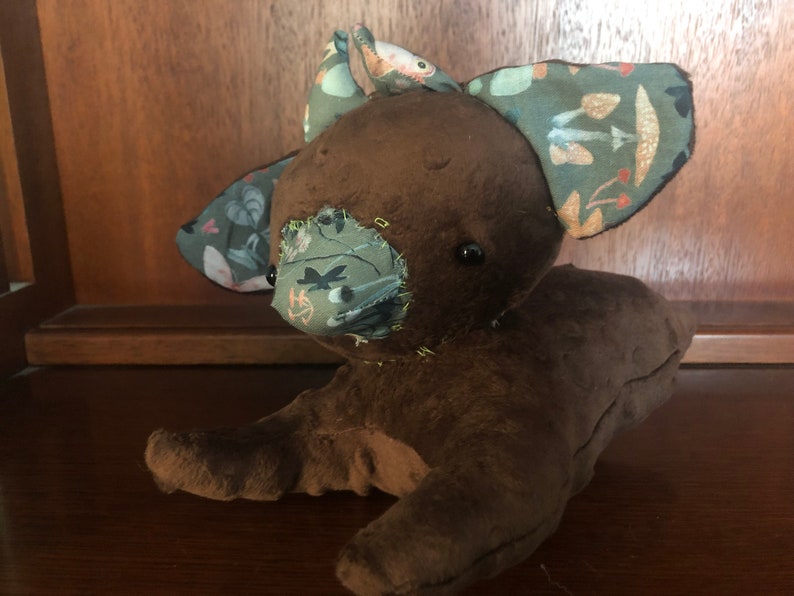 Op de afbeelding: Een bruin pluche speelgoed met een gestikt gezicht en stoffen oren. De oren zijn versierd met een groen, bruin en wit patroon. Het speelgoed heeft een gestikte neus en zwarte ogen.