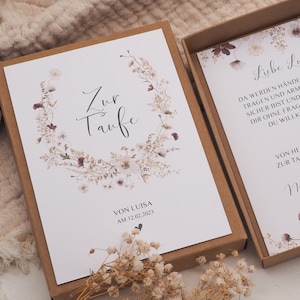 May include: A white card with a floral wreath and the text "Zur Taufe" in black calligraphy. The card is in a brown box with a smaller white card inside that reads "Liebe Luisa. Da werden H&auml;nde sein, die dich tragen und Arme, in denen du sicher bist und Menschen, die dir ohne Fragen zeigen, dass du willkommen bist. Von Herzen alles zur Taufe w&uuml;nscht, Nadita &"