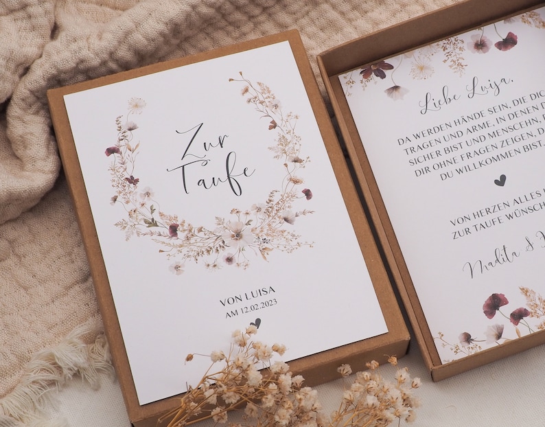May include: A white card with a floral wreath and the text "Zur Taufe" in black calligraphy. The card is in a brown box with a smaller white card inside that reads "Liebe Luisa. Da werden H&auml;nde sein, die dich tragen und Arme, in denen du sicher bist und Menschen, die dir ohne Fragen zeigen, dass du willkommen bist. Von Herzen alles zur Taufe w&uuml;nscht, Nadita &"