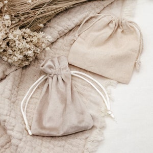 May include: Two small, beige fabric drawstring bags with white drawstrings. The bags are lying on a light brown textured surface.
