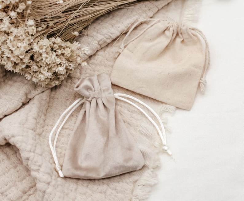 May include: Two small, beige fabric drawstring bags with white drawstrings. The bags are lying on a light brown textured surface.
