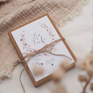 May include: A white card with a floral design and the text "Zur Taufe" in cursive. The card is tied with twine and placed in a brown box. The text "Wie schon, dass du geboren bist!" and "Von Luisa am 12.02.2023" is also visible on the card.