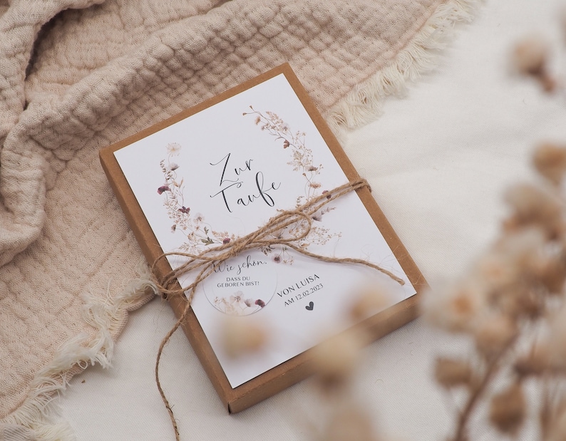 May include: A white card with a floral design and the text "Zur Taufe" in cursive. The card is tied with twine and placed in a brown box. The text "Wie schon, dass du geboren bist!" and "Von Luisa am 12.02.2023" is also visible on the card.