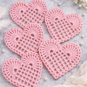 May include: Six pink crochet heart-shaped coasters. Each coaster features an openwork design with a central hole and a decorative border. The coasters are arranged on a light gray surface with pearls and feathers.
