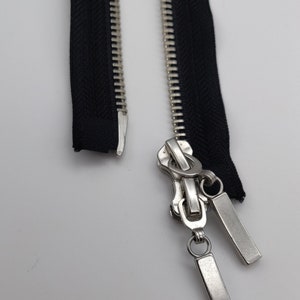 May include: A silver zipper with two black zipper tapes. The zipper has a pull tab with two metal loops.