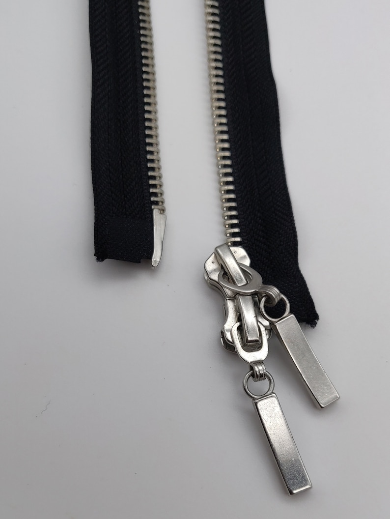 May include: A silver zipper with two black zipper tapes. The zipper has a pull tab with two metal loops.