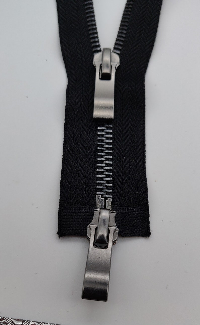 May include: A black zipper with a silver metal slider. The zipper is closed and the slider is in the middle of the zipper.