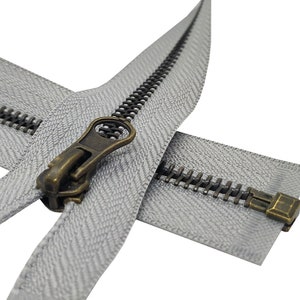 K&ouml;nnte beinhalten: Nahaufnahme eines silbernen Rei&szlig;verschlusses mit einem Messing-Schieber und -Zipper. Der Rei&szlig;verschluss ist ge&ouml;ffnet und die Z&auml;hne sind sichtbar. Der Rei&szlig;verschluss befindet sich auf einem wei&szlig;en Hintergrund.
