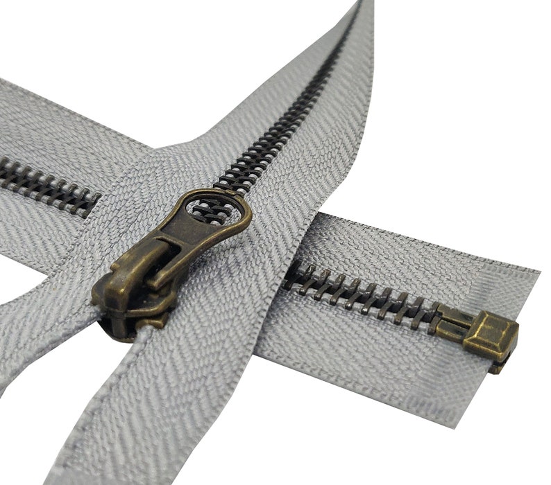 K&ouml;nnte beinhalten: Nahaufnahme eines silbernen Rei&szlig;verschlusses mit einem Messing-Schieber und -Zipper. Der Rei&szlig;verschluss ist ge&ouml;ffnet und die Z&auml;hne sind sichtbar. Der Rei&szlig;verschluss befindet sich auf einem wei&szlig;en Hintergrund.