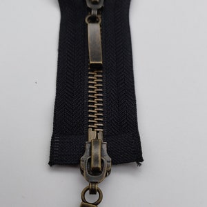 May include: A black zipper with a brass slider and pull tab. The zipper is closed and the slider is in the middle of the zipper.