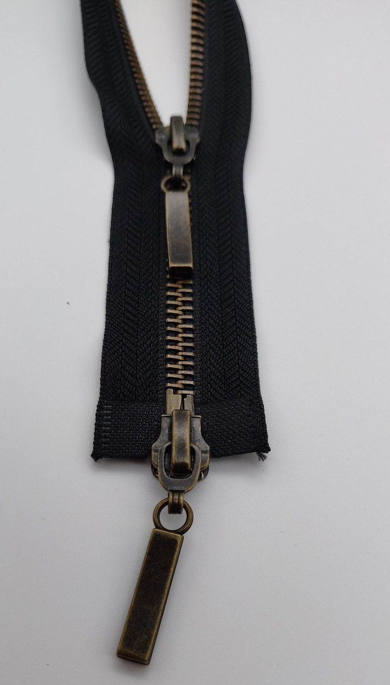 May include: A black zipper with a brass slider and pull tab. The zipper is closed and the slider is in the middle of the zipper.