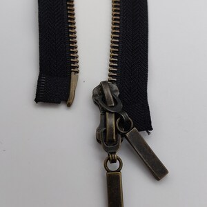 May include: A black zipper with a brass slider and two brass zipper pulls. The zipper is open and the slider is in the middle of the zipper.