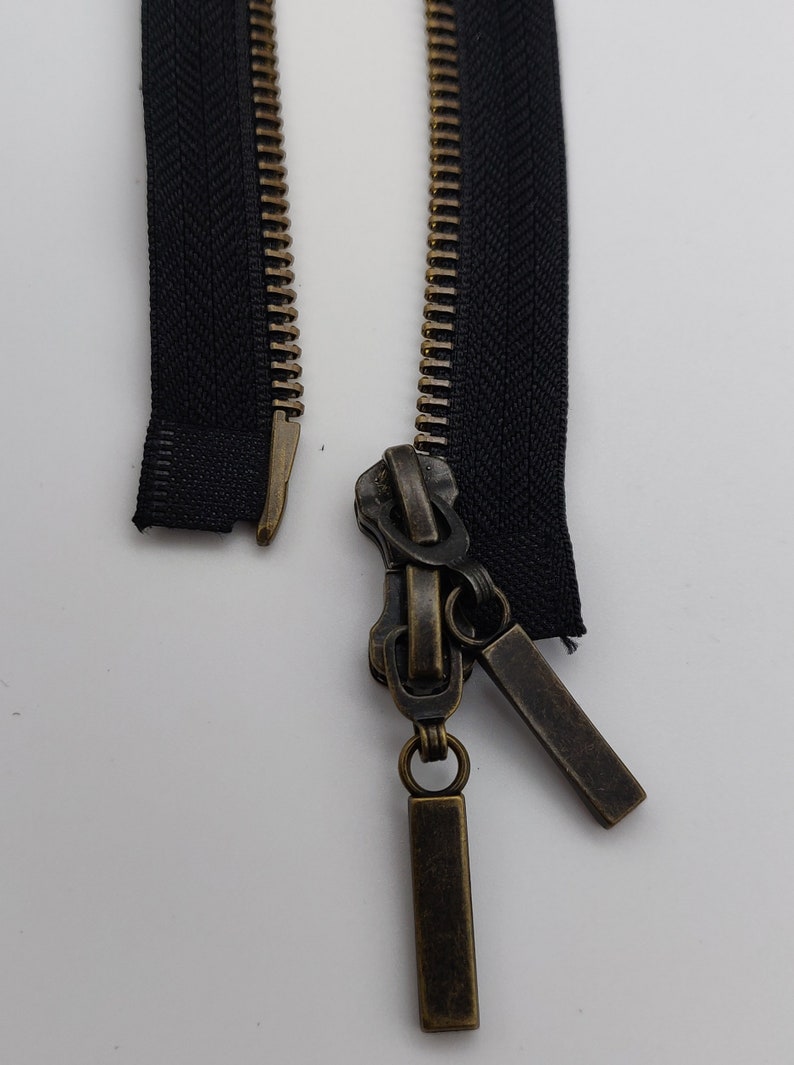 May include: A black zipper with a brass slider and two brass zipper pulls. The zipper is open and the slider is in the middle of the zipper.