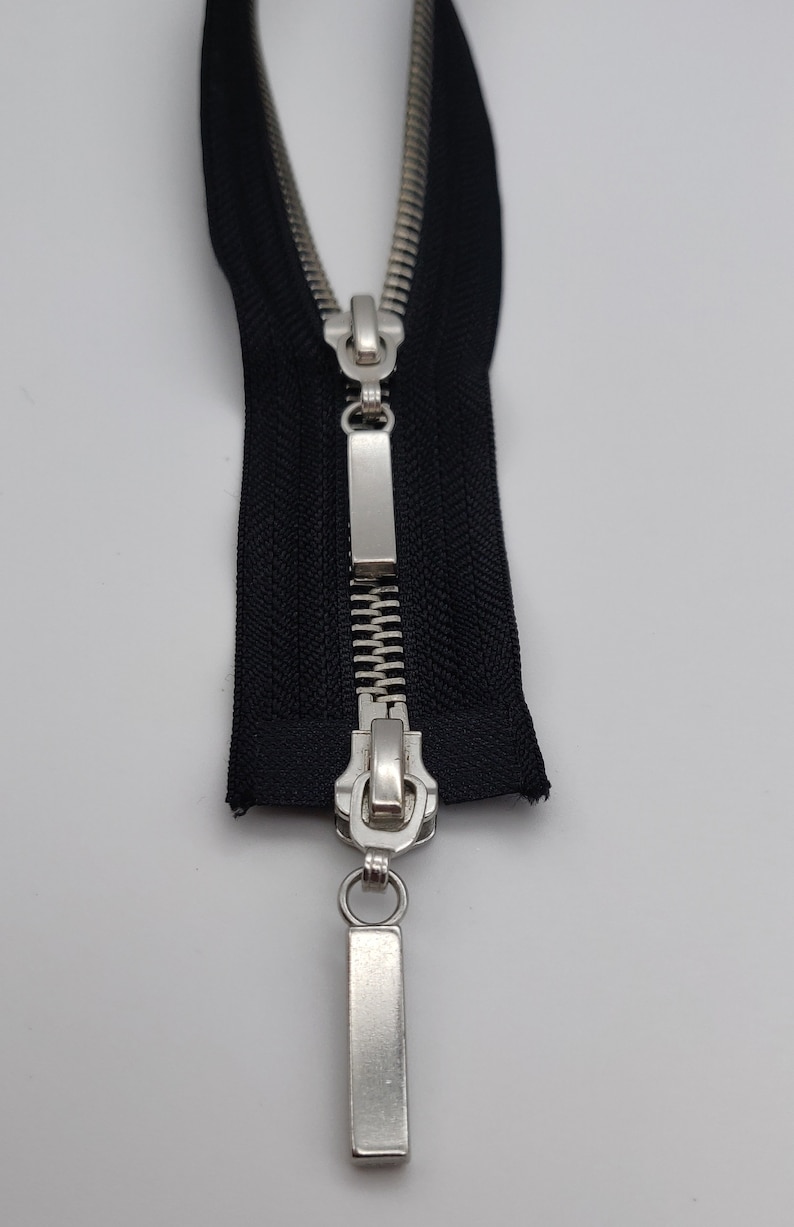 May include: A black zipper with a silver slider and pull tab. The zipper is open and the slider is in the middle of the zipper.