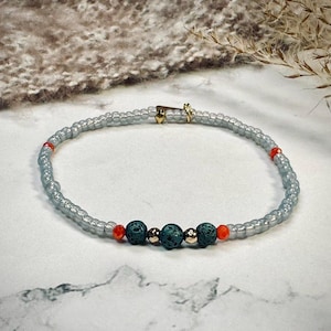 Könnte beinhalten: Ein zartes Armband aus hellblauen Perlen, akzentuiert mit drei dunkelgrünen Lavaperlen und kleinen orangefarbenen Perlen. Das Armband hat einen goldenen Verschluss.