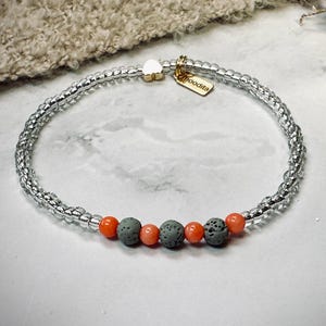 Könnte beinhalten: Ein Armband mit klaren, silberfarbenen Perlen und orangefarbenen und grauen Akzentperlen. Das Armband hat einen kleinen goldfarbenen Anhänger mit dem Wort "hoodita". Das Armband liegt auf einer weißen Oberfläche.