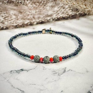 Könnte beinhalten: Ein zartes Armband aus kleinen, schillernden blauen Perlen und drei größeren grauen Lavasteinen mit roten Akzenten.