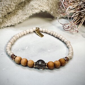 Könnte beinhalten: Ein Armband mit cremefarbenen Perlen, Holzperlen und einer Rauchquarzperle. Das Armband hat goldfarbene Akzente und ein kleines goldenes Etikett. Das Armband ist neutralfarben und für den täglichen Gebrauch geeignet.
