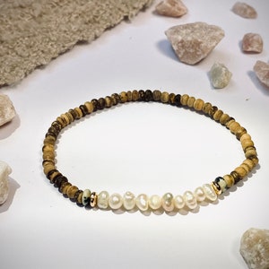 Könnte beinhalten: Perlenarmband mit einer Mischung aus Naturholz und cremefarbenen Süßwasserperlen. Das Armband hat kleine, runde Holzperlen in Braun- und Beigetönen, mit einem Abschnitt aus weißen Perlen. Goldfarbene Akzente sind ebenfalls vorhanden.