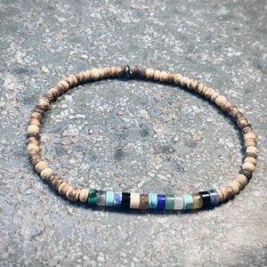 Könnte beinhalten: Ein Armband mit kleinen, runden, hellbraunen Perlen und farbigen Akzentperlen. Die Akzentperlen umfassen Schwarz, Blau, Grün und Weiß und setzen farbliche Akzente. Das Armband hat einen Umfang von etwa 18 cm.