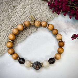 Op de afbeelding: Een kralenarmband met lichtbruine houten kralen, donkerbruine gefacetteerde kralen en twee gladde off-white kralen. De armband bevat kleine goudkleurige metalen tussenstukjes en een klein goudkleurig metalen label met de inscriptie moodita.