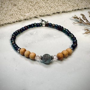 Könnte beinhalten: Ein Armband mit dunklen, irisierenden Perlen, hellbraunen Holzperlen und einer zentralen grauen Steinperle. Das Armband ist mit silberfarbenen Metallperlen und klaren Kristallperlen akzentuiert.
