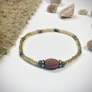 Könnte beinhalten: Ein zartes Armband aus kleinen goldenen, braunen und türkisfarbenen Perlen. Eine größere, ovale, braune Lavasteinperle ist der Mittelpunkt des Armbands.