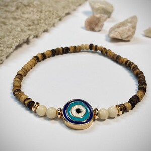 Könnte beinhalten: Ein Armband mit Holzperlen und einem bösen Blick-Anhänger. Das Armband besteht aus kleinen, hellbraunen und dunkelbraunen Holzperlen. Der Anhänger des bösen Blicks hat ein blaues und weißes Design mit goldfarbenen Akzenten.