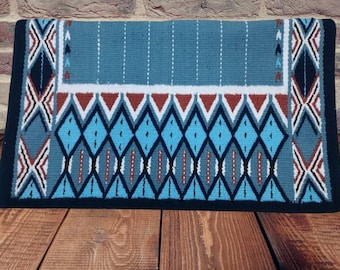 May include: A rectangular turquoise saddle pad with a black border. The pad features a geometric pattern with white, red, and black accents. The design includes diamonds, chevrons, and vertical stripes, creating a Southwestern aesthetic.
