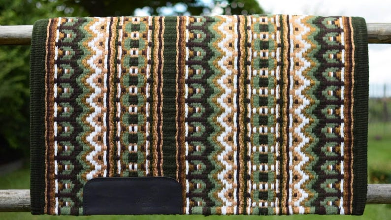 May include: A woven saddle pad with a geometric pattern in shades of green, brown, and white. The pad has a dark green border and a black leather patch at the bottom. The pad is displayed on a wooden fence.