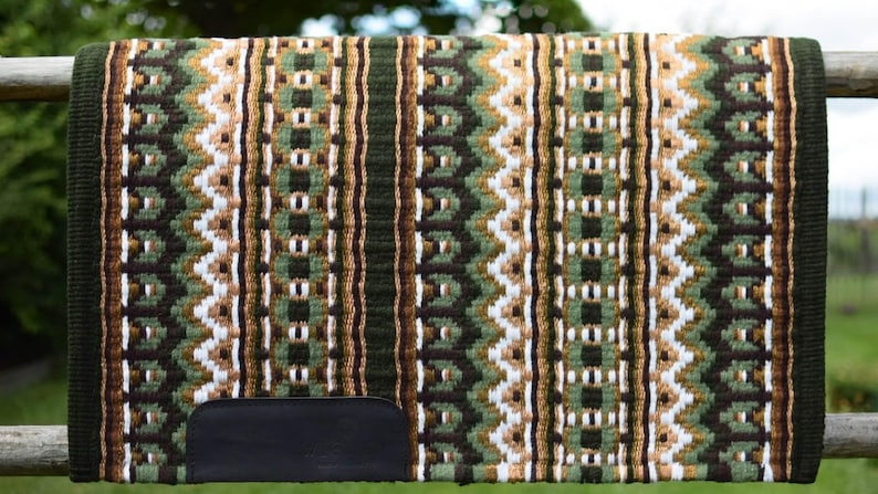 May include: A woven saddle pad with a geometric pattern in shades of green, brown, and white. The pad has a dark green border and a black leather patch at the bottom. The pad is displayed on a wooden fence.