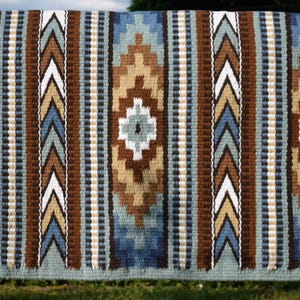 Könnte beinhalten: Eine gewebte Satteldecke mit einem geometrischen Muster in Blau-, Braun-, Beige- und Weißtönen. Das Design zeigt Diamanten- und Pfeil-Motive, die von einem hellgrünen Rand umgeben sind. Die Decke wird im Freien gezeigt.