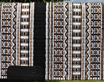 Puede incluir: Una almohadilla rectangular para silla de montar de caballo con una base negra y un patrón geométrico repetido en tonos marrones, blancos y grises. La almohadilla tiene un parche de cuero negro en la esquina inferior izquierda. La almohadilla se muestra en una valla de madera.